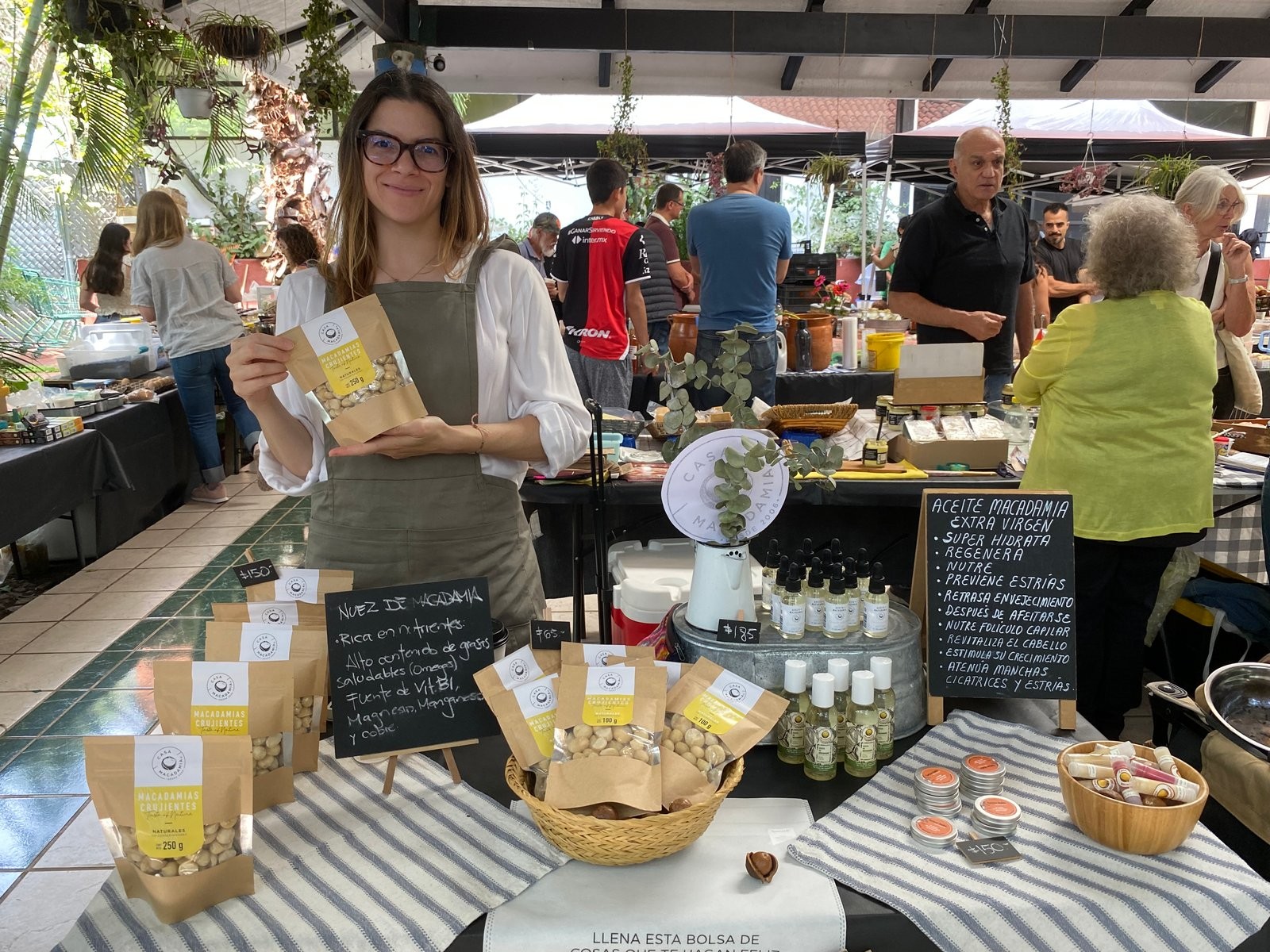 Responsable de Casa Macadamia en el puesto, mostrando una bolsa de macadamias crujientes junto a frascos de aceite y un pizarrón de beneficios