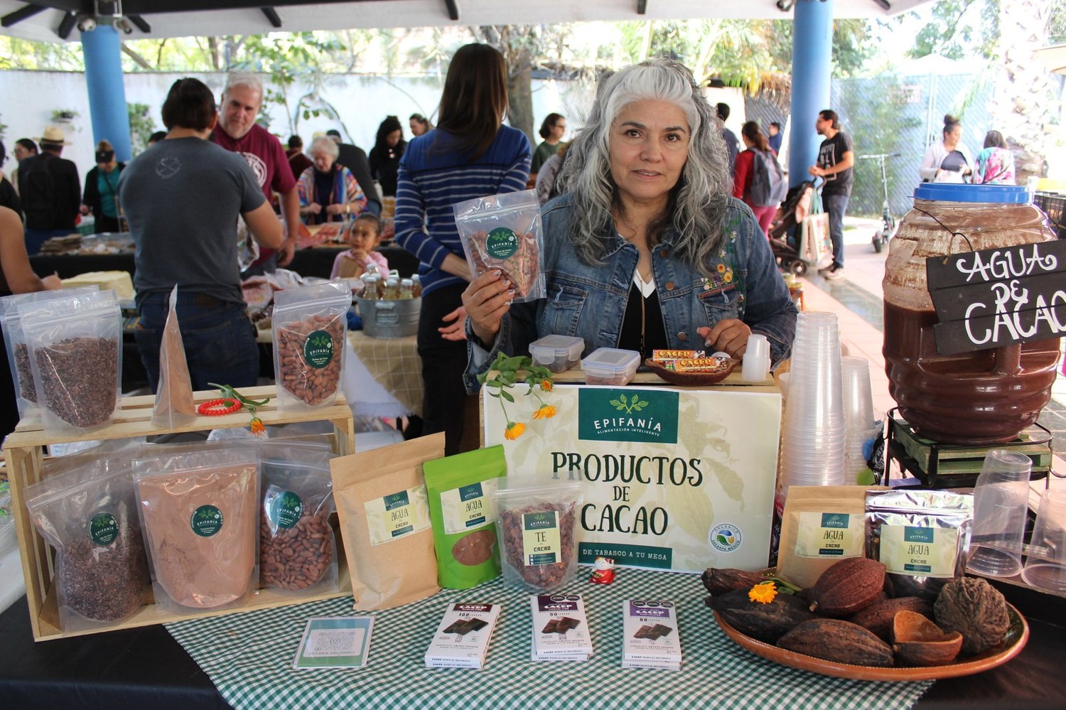 Puesto de Epifanía con vainas de cacao fresco, bolsas de grano y polvo, y el garrafón de agua de cacao de fondo