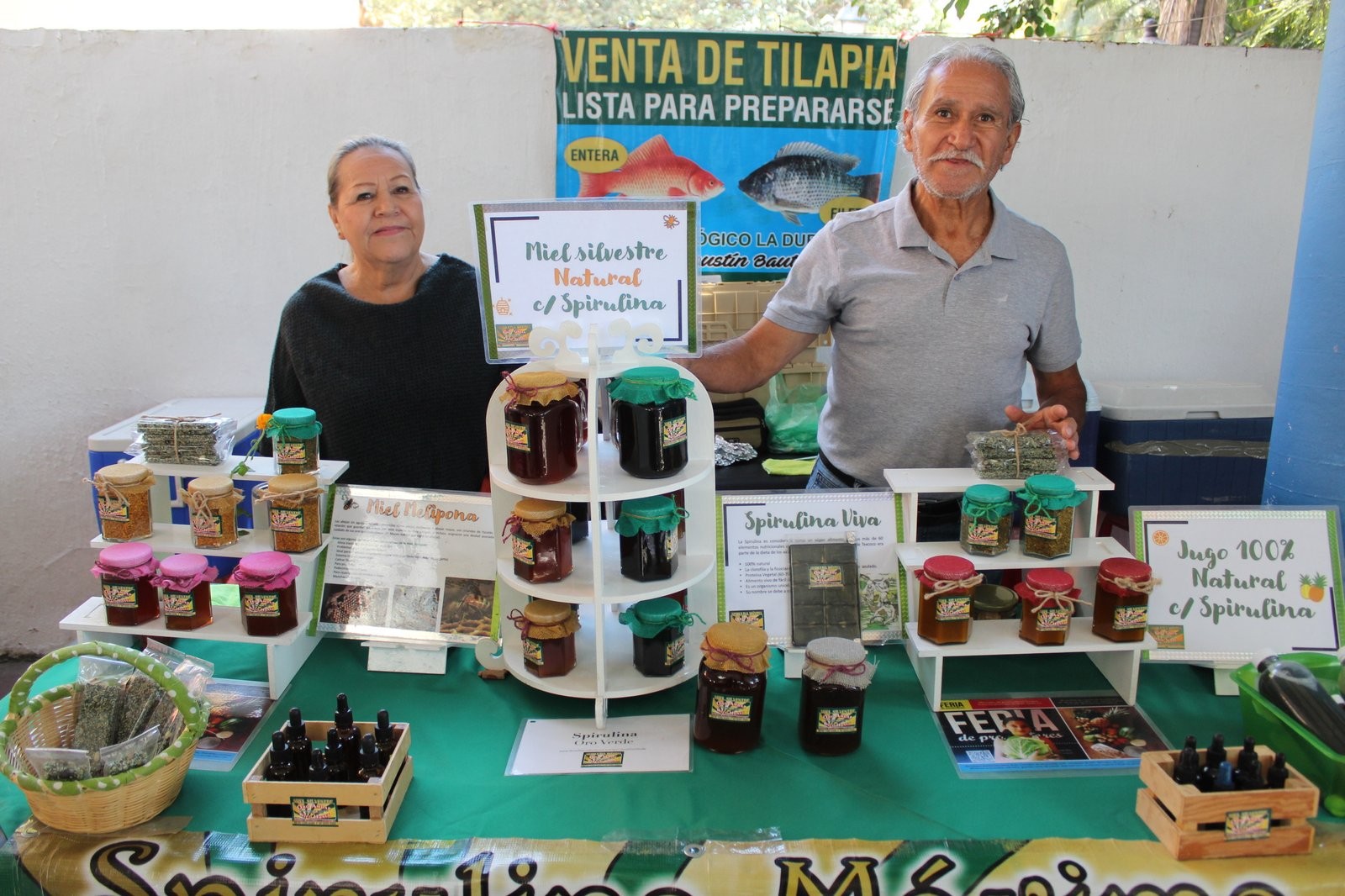 Puesto de Espirulina Oro Verde con frascos de miel silvestre y miel melipona, bajo un cartel de venta de tilapia