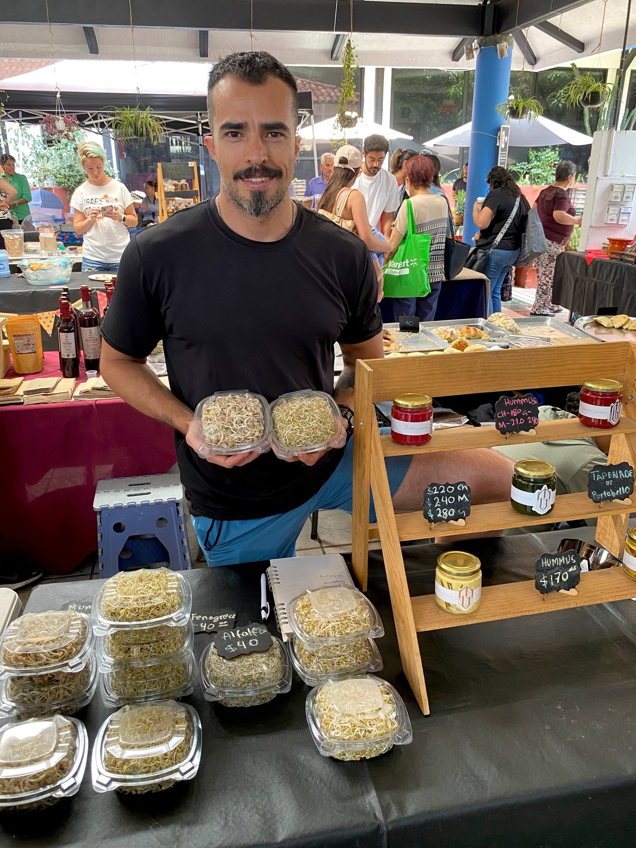 Productor de Magick Garden sosteniendo charolas de germinados, con el estante de hummus y tapenade al fondo