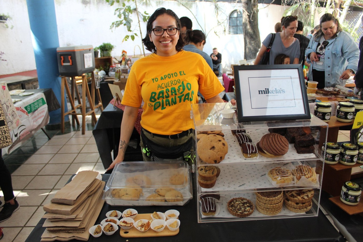 Fundadora de Mikela's en su puesto, junto a la vitrina con galletas, conchas veganas, brownies y tartaletas, y frascos de mermelada