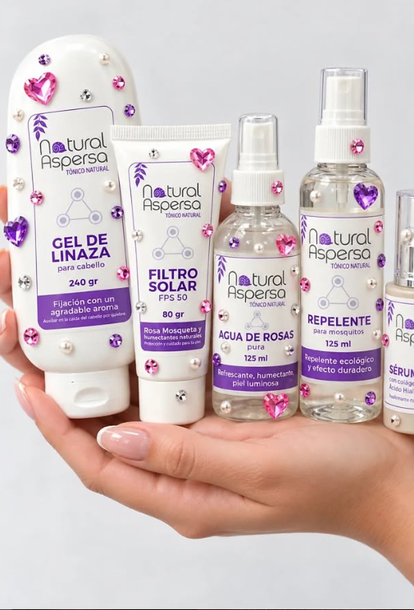 Mano sosteniendo seis productos de Natural Aspersa: gel de linaza, filtro solar, agua de rosas, repelente y serum