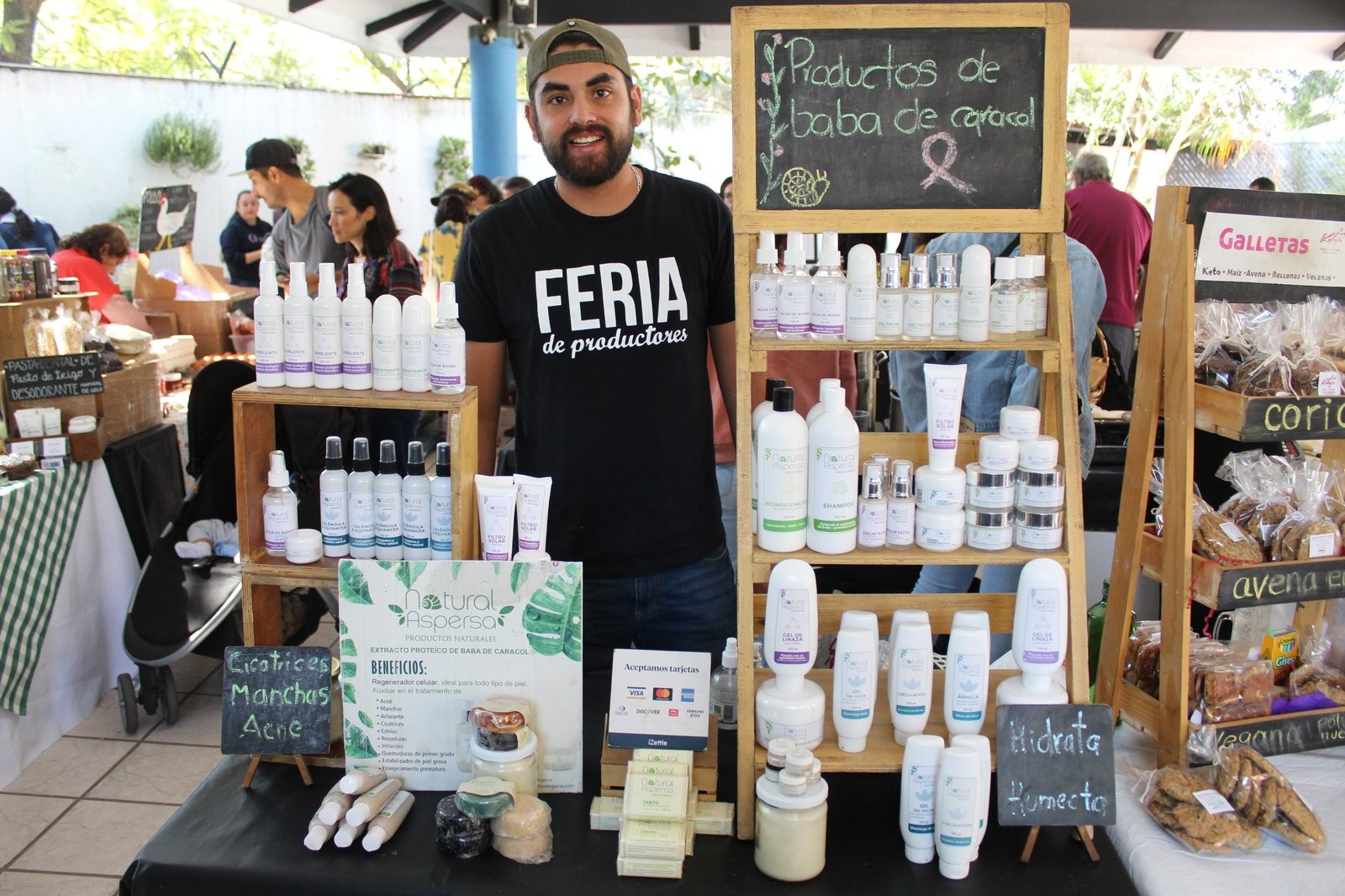 Carlos Eduardo Gelaviz en el puesto de Natural Aspersa, con el pizarrón 'Productos de baba de caracol' y un estante de frascos