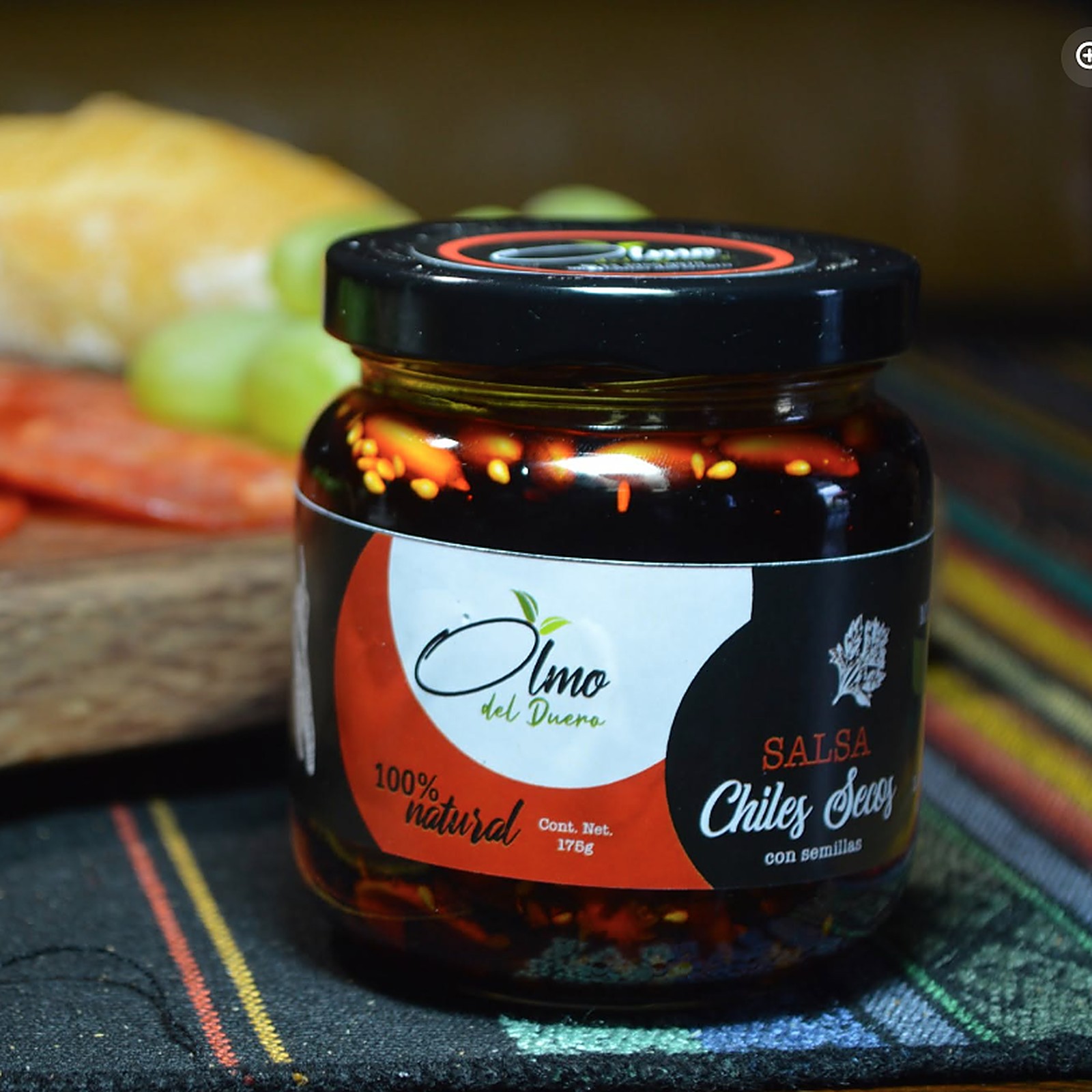 Frasco de salsa de chiles secos con semillas de Olmo del Duero, sobre una mesa con quesos y verduras de fondo