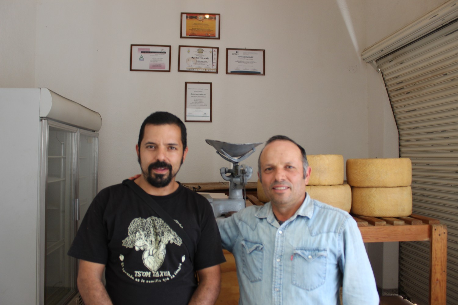 Dos integrantes de la familia junto a ruedas de queso en tarima de madera, con diplomas y reconocimientos en la pared