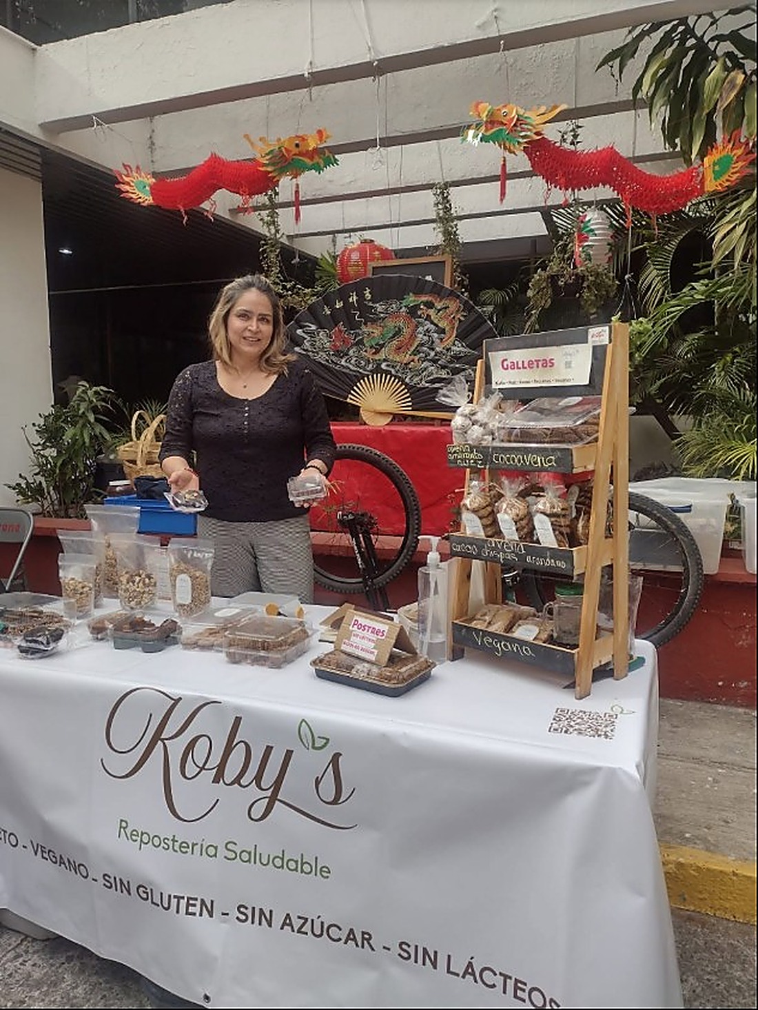 Irma en el puesto de Koby's, con un mantel que dice KETO - VEGANO - SIN GLUTEN - SIN AZÚCAR - SIN LÁCTEOS, junto a un estante de galletas empacadas